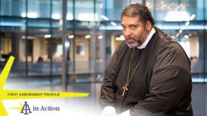A photo showing William Barber in a glass-enclosed room, looking sidelong into the camera and wearing a large cross, with text overlay reading First Amendment Profile: 1A in Action
