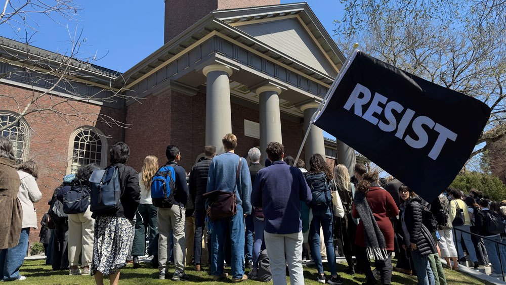 An image of a group of people standing in front of a brick building, with one person holding a large sign reading "resist"