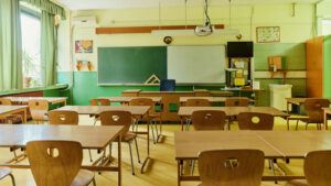 A classroom with wooden chairs and desks, and a green chalkboard, sits empty