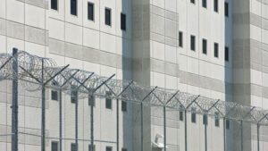 A close-up image of a prison building with grey walls and small windows, and fencing in the front