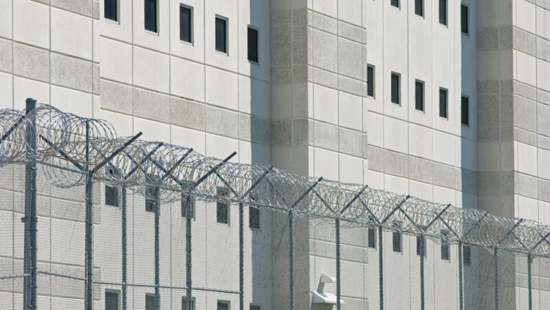 A close-up image of a prison building with grey walls and small windows, and fencing in the front
