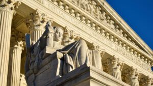 A close-up view of the facade of the Supreme Court building, with the front face of the building and a statue in the foreground and a clear blue sky in the background