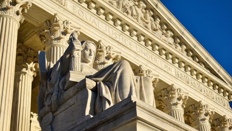 A close-up view of the facade of the Supreme Court building, with the front face of the building and a statue in the foreground and a clear blue sky in the background