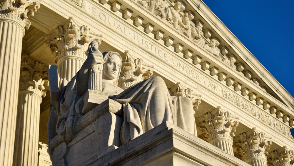 A close-up view of the facade of the Supreme Court building, with the front face of the building and a statue in the foreground and a clear blue sky in the background