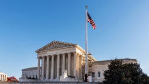 A front view of the U.S. Supreme Court building against a clear blue sky, with an American flag flying in front