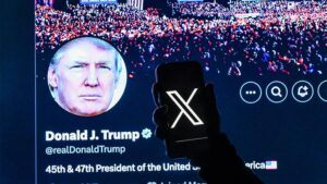 An illustrative photo of a woman holding a smartphone displaying the X logo, with the cover page of President Donald Trump's X account visible on a computer screen in the background