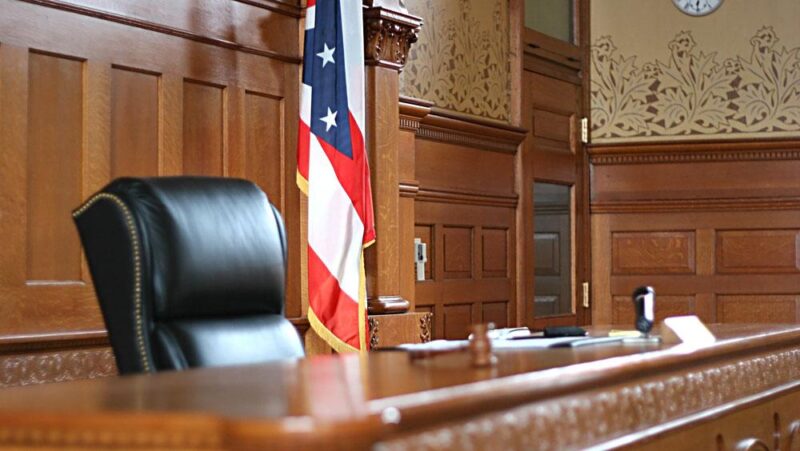 A photo of the interior of a courtroom, with wood paneling lining the room, a commanding black leather chair in the foreground, and a partial view of the U.S. flag in the background