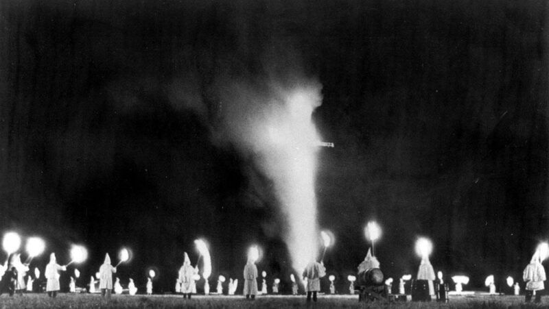 A black and white image of a cross burning at a Ku Klux Klan rally, with Klan members in white robes holding torches encircle a burning cross