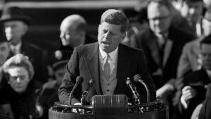 Black and white image of JFK speaking at a podium with a crowd standing behind him