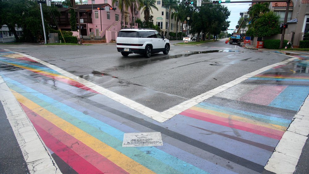 An image showing two intersecting rainbow crosswalks, with a white SUV driving nearby