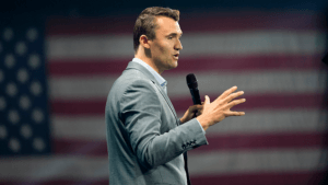 Charlie Kirk speaks in a microphone with an American flag in the background
