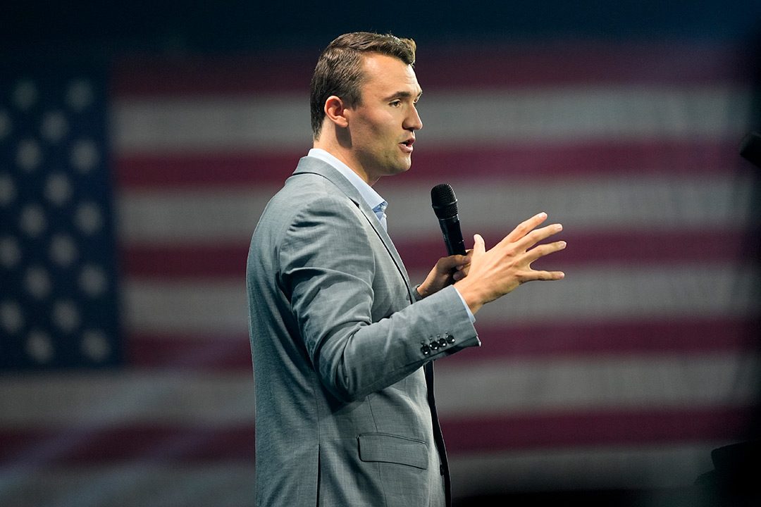 Charlie Kirk speaks in a microphone with an American flag in the background
