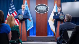 Pete Hegseth and Dan Caine lead a press conference at the Pentagon, standing in front of two podiums with the Pentagon seal behind them