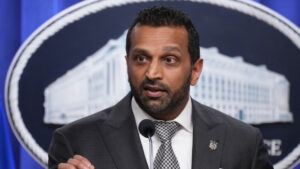 Kash Patel, wearing a suit and tie, speaks as he stands in front of a sign depicting an image of the Department of Justice building, with the agency name written below