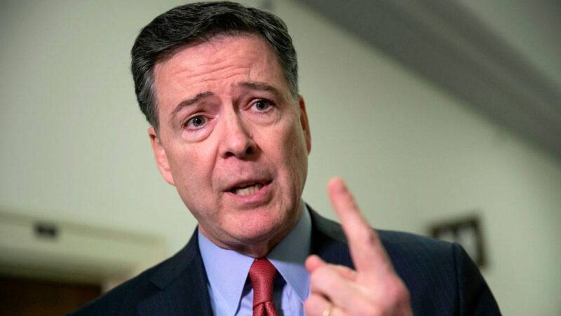 James Comey, wearing a suit and tie, raises his hand as he speaks