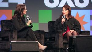 Selena Gomez, wearing a black jacket over a red dress, sits to the right of Dr. Jessica Stern. Both are holding mics and speaking on a stage at a convention center