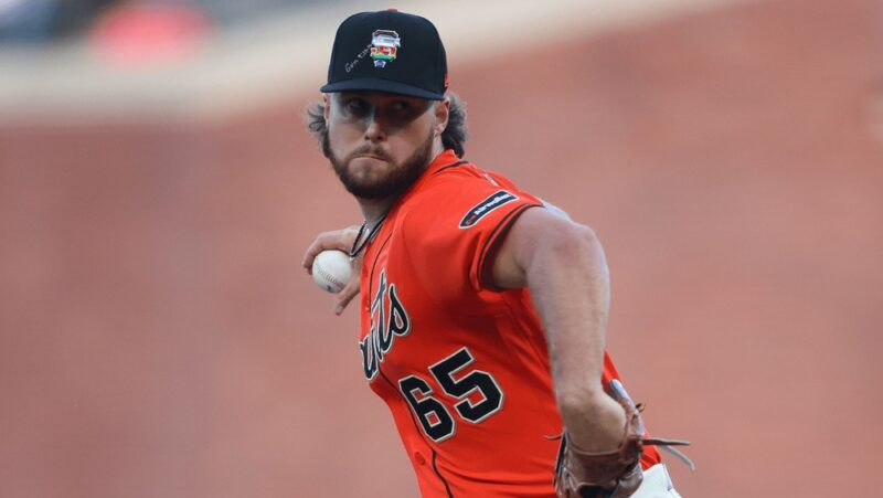 A baseball player wearing a bright-orange jersey prepares to throw a ball. He wears a hat with the rainbow version of the team logo, next to handwritten text that references a Genesis Bible verse.